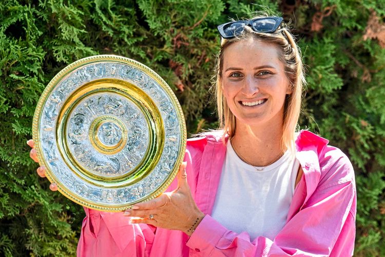 Eine lächelnde Frau in einem pinken Hemd hält eine silbrig-goldene Trophäe mit detaillierten Verzierungen. Im Hintergrund sind grüne Büsche zu sehen.