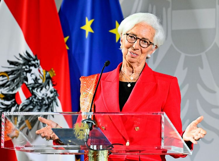 Eine Frau mit weißem Haar, Brille und rotem Blazer steht an einem Rednerpult mit Mikrofon. Im Hintergrund sind die Flaggen von Österreich und der Europäischen Union zu sehen.