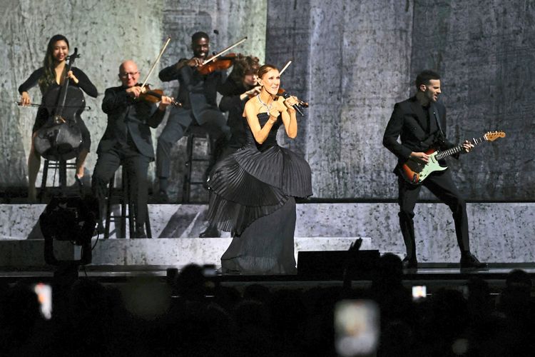 Eine Sängerin in einem schwarzen Kleid steht auf einer Bühne und singt in ein Mikrofon. Im Hintergrund spielen Musiker auf Streichinstrumenten und eine E-Gitarre. Die Szene ist stimmungsvoll beleuchtet, und das Publikum ist im Vordergrund teilweise sichtbar.