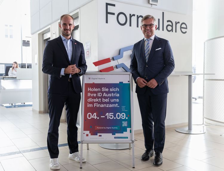 Die beiden Herren stehen im Anzug im Finanzministerium vor einer Tafel mit der Aufschrift: Holen Sie sich Ihre ID Austria direkt bei uns im Finanzamt. Vierter bis 15. September 2023. 