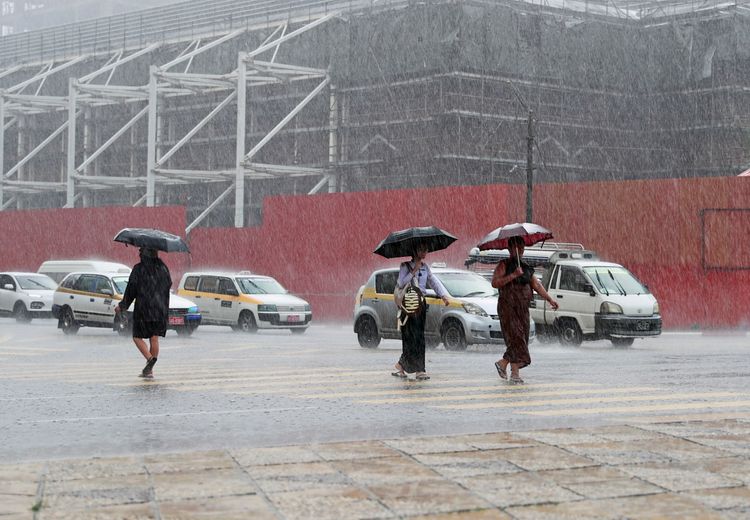 Drei Personen gehen bei starkem Regen mit Regenschirmen über einen Zebrastreifen in Yangon, Myanmar. Im Hintergrund sind parkende Autos und eine Baustelle mit einem Gerüst zu sehen.