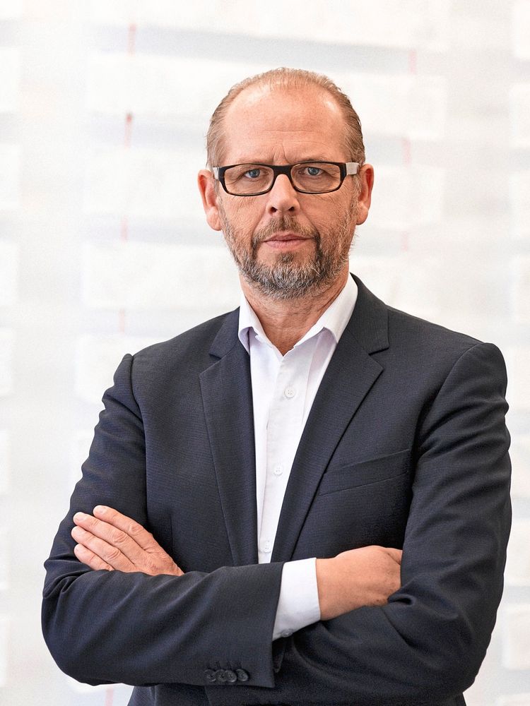 Ein Mann mit Brille, Bart und kurzen Haaren trägt ein dunkles Sakko mit weißem Hemd. Er verschränkt die Arme vor der Brust und blickt in die Kamera. Der Hintergrund ist hell und unscharf.