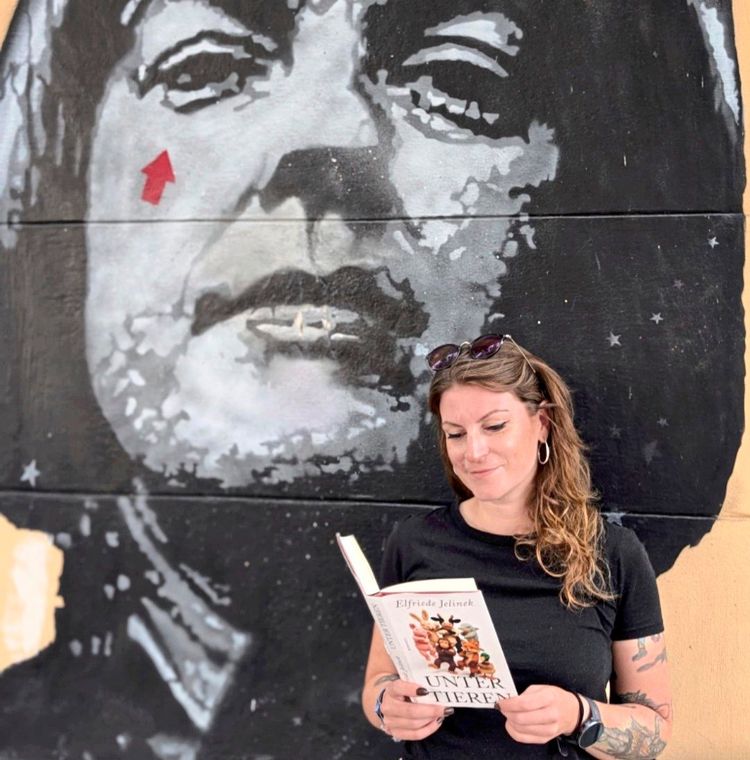 Eine Frau mit langen, braunen Haaren steht vor einer Wand mit einem großen Schwarz-Weiß-Graffiti eines Gesichts. Sie hält ein Buch mit dem Titel 