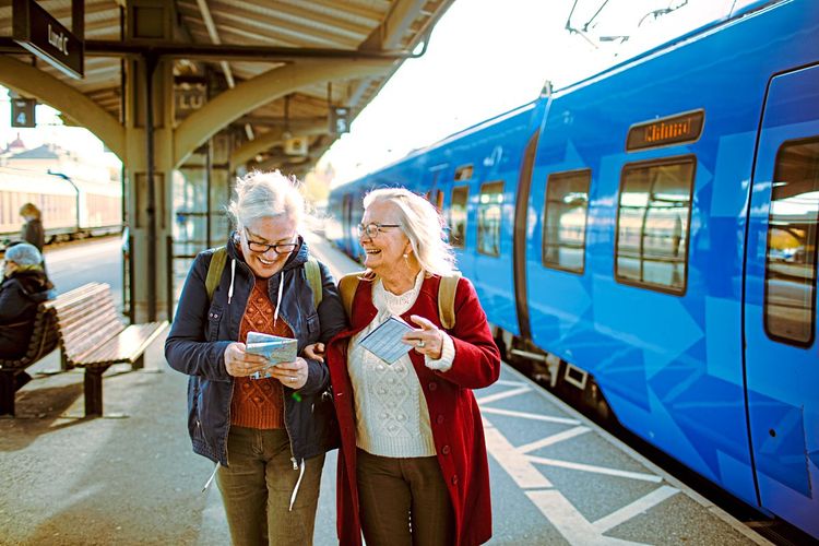 Zwei ältere Damen am Bahnsteig, lachend und mit Fahrplänen beschäftigt.