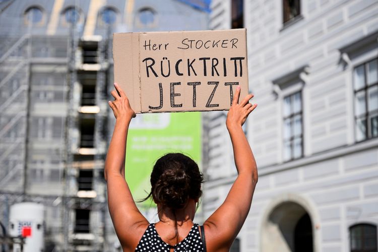 Eine Frau hält ein Schild mit der Aufschrift „Herr Stocker Rücktritt Jetzt“ in die Luft, während sie an einer Demonstration in einer städtischen Umgebung teilnimmt. Im Hintergrund sind Gebäude und Baugerüste zu sehen.