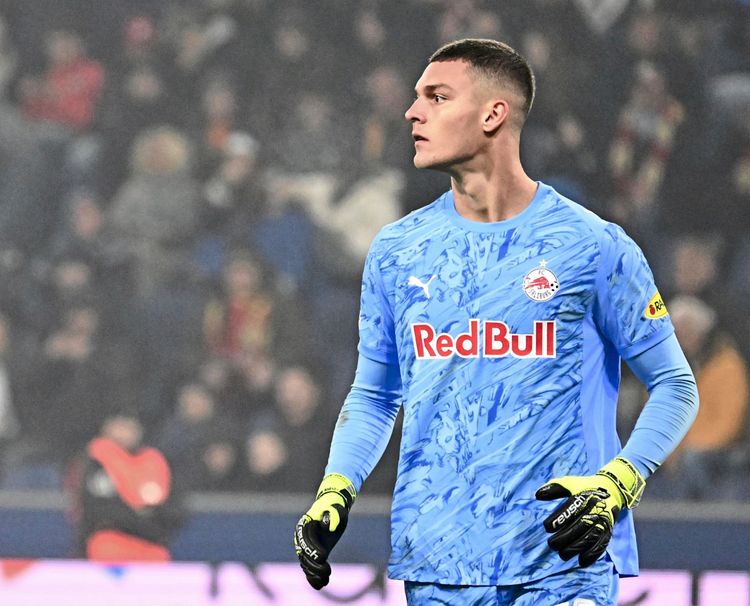 Ein Fußballspieler im blauen Torwarttrikot von Red Bull Salzburg steht auf dem Spielfeld, mit Blick zur Seite. Im Hintergrund unscharf erkennbar sind Zuschauer auf den Tribünen.