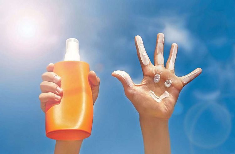 Eine Hand hält vor einem blauen Himmel eine orange Flasche Sonnencreme hoch, daneben eine Handfläche, auf die mit Sonnencreme ein Smiley gemalt ist