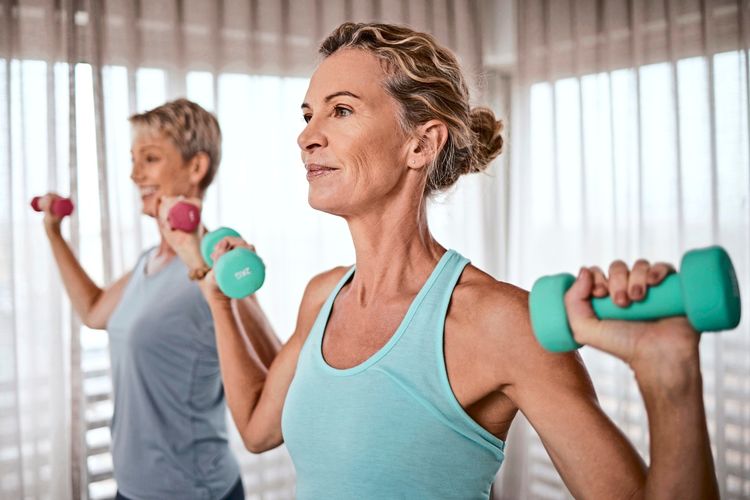 Zwei Frauen trainieren mit Kurzhanteln in einem hellen Raum. Sie tragen Sportkleidung und wirken konzentriert. Im Hintergrund sind Vorhänge zu sehen.