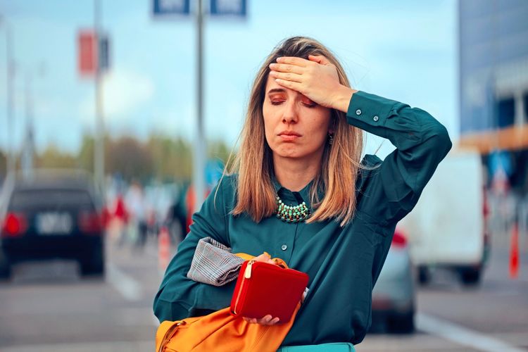 Eine Frau trägt eine grüne Bluse, hält eine gelbe Tasche sowie eine rote Brieftasche und wirkt beunruhigt auf einer belebten Straße. Im Hintergrund sind unscharfe Autos und Passanten zu sehen.