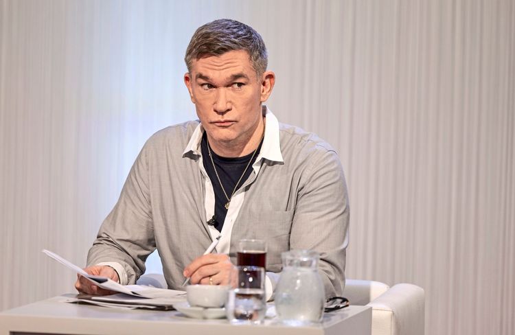 Ein Mann mit grauem Hemd und offenem Kragen sitzt an einem Tisch, hält Papiere in der Hand und schaut nachdenklich zur Seite. Vor ihm stehen ein Glas Wasser, eine Karaffe und eine Tasse. Der Hintergrund ist hell und schlicht.