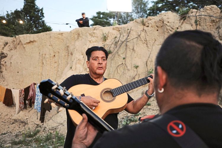 Ein Mann spielt eine Akustikgitarre vor einer sandigen Felswand im Freien. Im Vordergrund sieht man den Rücken einer weiteren Person mit einer Gitarre. Im Hintergrund hängen bunte Stoffe an einer Leine, und ein Mann steht erhöht auf der Klippe. Die Szene wirkt lebendig und kulturell geprägt.