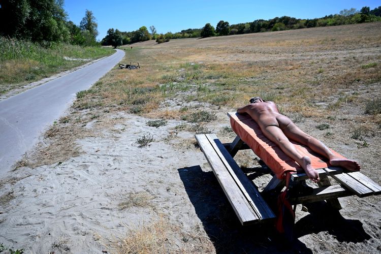 Eine Person liegt nackt auf einem orangefarbenen Handtuch auf einem Holztisch in einer offenen Landschaft unter blauem Himmel. Ein Fahrrad liegt auf einem Weg links im Hintergrund. Die Umgebung ist trocken und grasbewachsen.