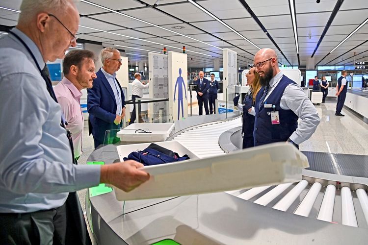Ein moderner Sicherheitsbereich am Flughafen mit mehreren Sicherheitsmitarbeitern und Reisenden, die an einer Handgepäckkontrolle teilnehmen. Ein Förderband mit Scannern ist sichtbar, und Gegenstände werden sorgfältig überprüft. Die Szene stammt aus der Eröffnung des Terminals 1-Pier am Flughafen Franz Josef Strauß in München am 13. April 2026.