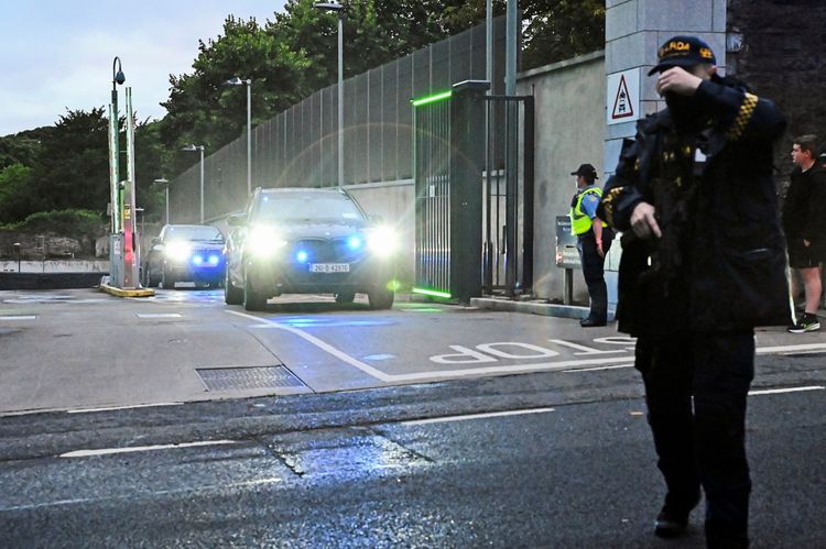 Ein schwer bewachtes Fahrzeug mit eingeschaltetem Blaulicht verlässt ein umzäuntes Gelände durch ein geöffnetes Tor. Bewaffnete Sicherheitskräfte und Polizisten sind im Vorder- und Hintergrund sichtbar. Mehrere Personen beobachten die Szene.