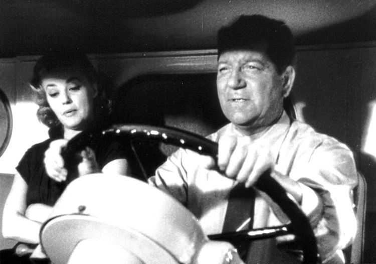 Ein Schwarz-Weiß-Bild zeigt eine Frau und einen Mann in einem Fahrzeug. Der Mann sitzt am Steuer, trägt ein Hemd und hält das Lenkrad. Die Frau schaut auf ihre Hand und scheint etwas zu zeigen oder zu erklären.