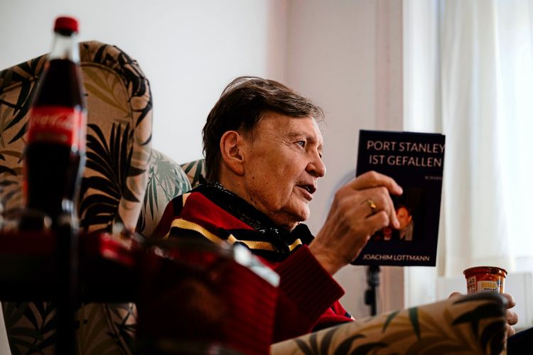Ein älterer Mann sitzt in einem gemusterten Sessel in einem Zimmer mit hellem Tageslicht. Er hält ein Buch mit dem Titel „Port Stanley ist gefallen“ von Joachim Lottmann in der Hand. Auf einem Beistelltisch ist eine Cola-Flasche sichtbar.