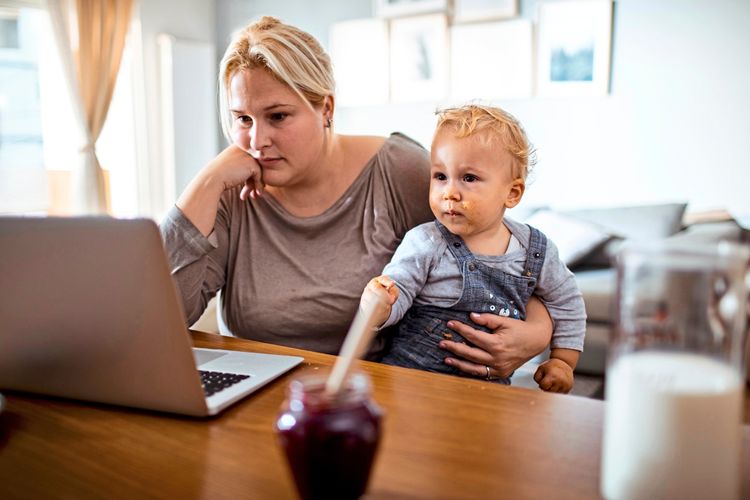 Eine junge Frau sitzt nachdenklich vor einem Laptop und hält ein Kleinkind auf dem Schoß. Das Kind trägt Latzhosen und hat Essensreste im Gesicht. Auf dem Tisch stehen ein Glas Milch und ein Glas mit einem Löffel.