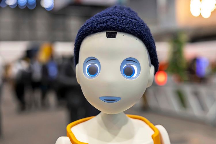 Ein sozialer Roboter namens Navel von Navel Robotics, ausgestattet mit künstlicher Intelligenz, trägt eine blaue Mütze und ein orangefarbenes Accessoire, aufgenommen während des AI for Good Global Summit im Palexpo in Genf, Schweiz. Im Hintergrund sind unscharfe Besucher und Veranstaltungsdetails zu sehen.