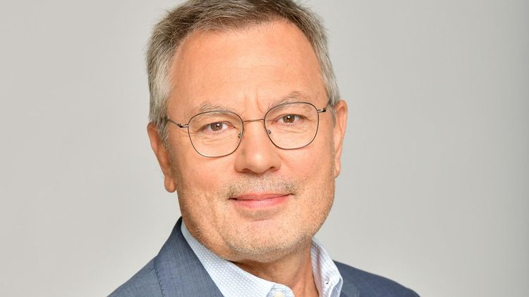 Mann mit kurzen Grauen Haaren, Metallbrille, blaugrauem Sakko und hellem Hemd lächelt in die Kamera (Porträt).
