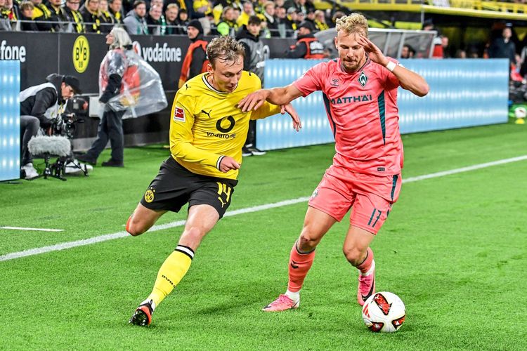Zwei Fußballspieler im Zweikampf während eines Spiels im Signal Iduna Park, Dortmund. Der Spieler links im gelben Trikot von Borussia Dortmund tritt gegen den Spieler rechts im pinken Trikot von Werder Bremen an. Im Hintergrund sind Zuschauer und Stadionwerbung zu sehen.‘