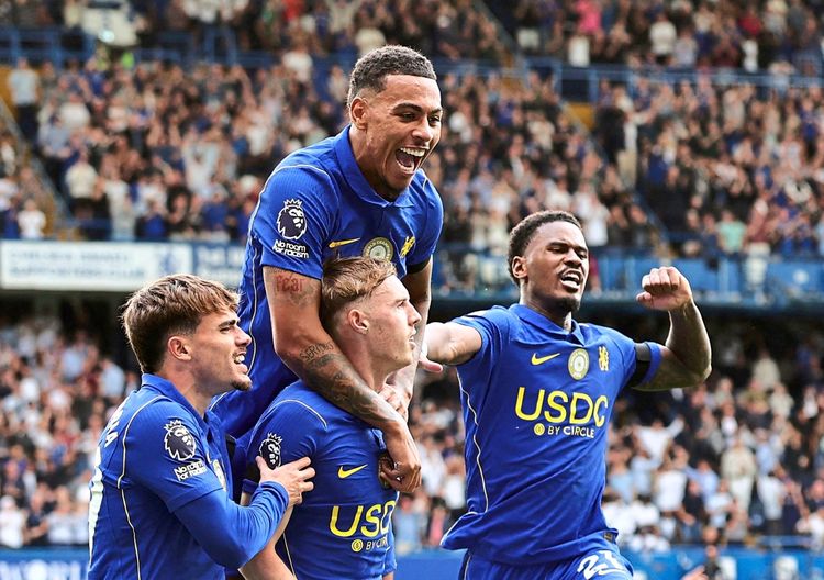 Vier Fußballspieler im blauen Chelsea-Trikot feiern ein Tor während eines Spiels im Stadion. Einer von ihnen wird von einem anderen hochgehoben, während die anderen jubeln und ihre Freude ausdrücken. Im Hintergrund sind jubelnde Fans auf den Tribünen zu sehen.