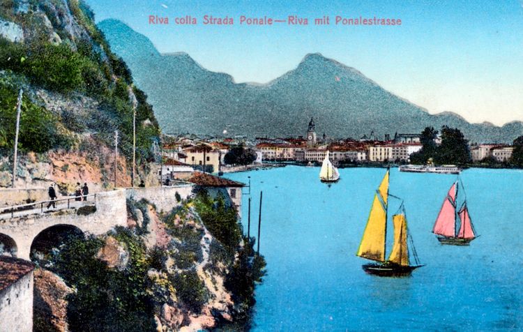 Ansichtskarte von Riva del Garda am Gardasee, Italien, um 1905. Im Vordergrund ist die Ponalestraße zu sehen, die entlang eines steilen Abhangs verläuft. Im See segeln bunte Boote, und im Hintergrund liegt die Stadt vor einer Bergkulisse.