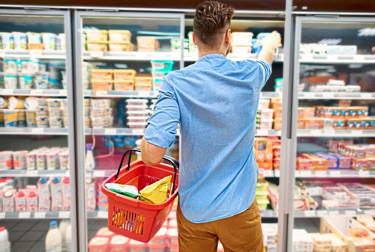 Ein Mann steht mit seinem Einkaufskorb vor einer Kühlvitrine und greift nach einem Produkt. 