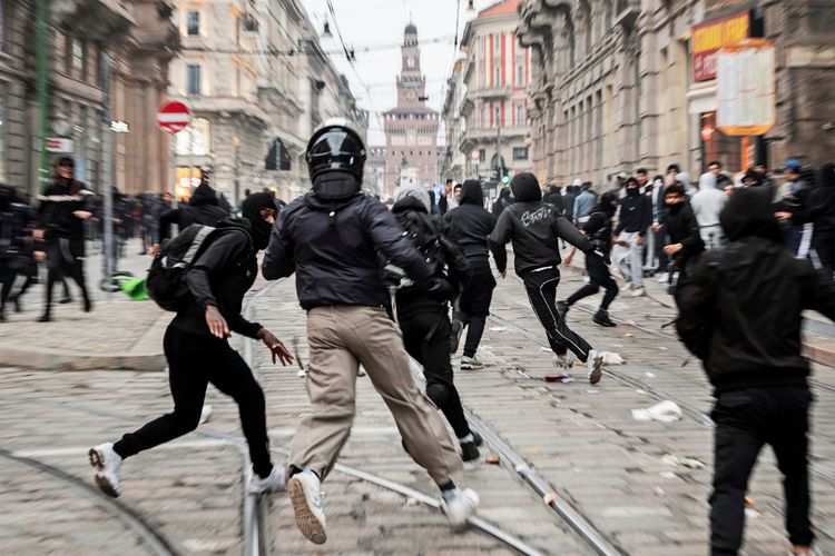 Straßenszene in Mailand während einer Demonstration gegen die hohen Lebenshaltungskosten. Menschen in Masken und dunkler Kleidung laufen und bewegen sich in verschiedene Richtungen, während Müll auf der gepflasterten Straße verstreut liegt. Im Hintergrund sind historische Gebäude zu sehen.