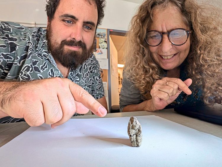 Zwei Personen zeigen auf ein kleines, ungewöhnlich geformtes, steinzeitliches Artefakt, das auf einem weißen Papier liegt. Im Hintergrund ist ein Büro mit Notizen und einem offenen Türrahmen zu sehen.
