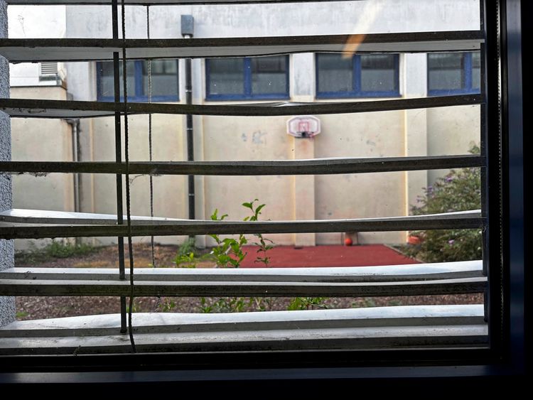 Blick durch vergittertes Fenster auf einen kleinen Hof mit rotem Bodenbelag, Basketballkorb an einer beigen Wand und Pflanzen im Vordergrund.