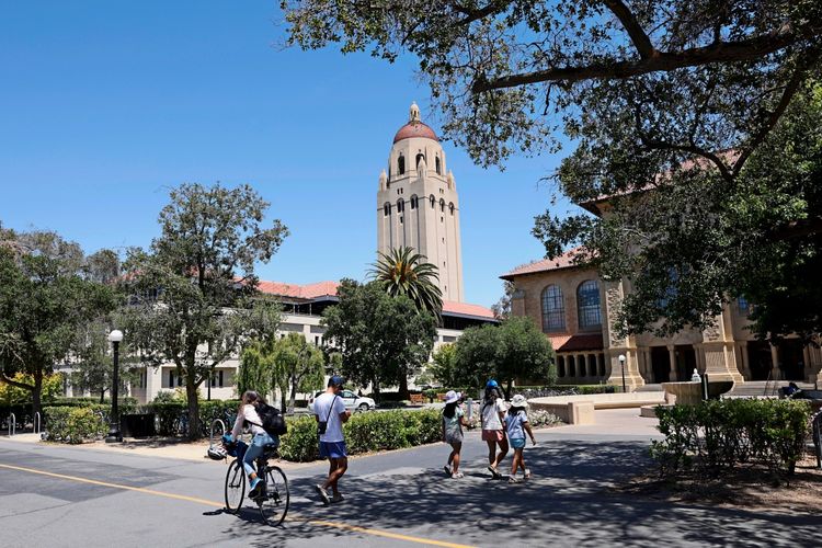 Menschen spazieren und fahren mit dem Fahrrad über den Campus der Stanford University in Stanford, Kalifornien, an einem sonnigen Tag. Im Hintergrund sind Gebäude der Universität und der markante Turm zu sehen, umgeben von Bäumen.