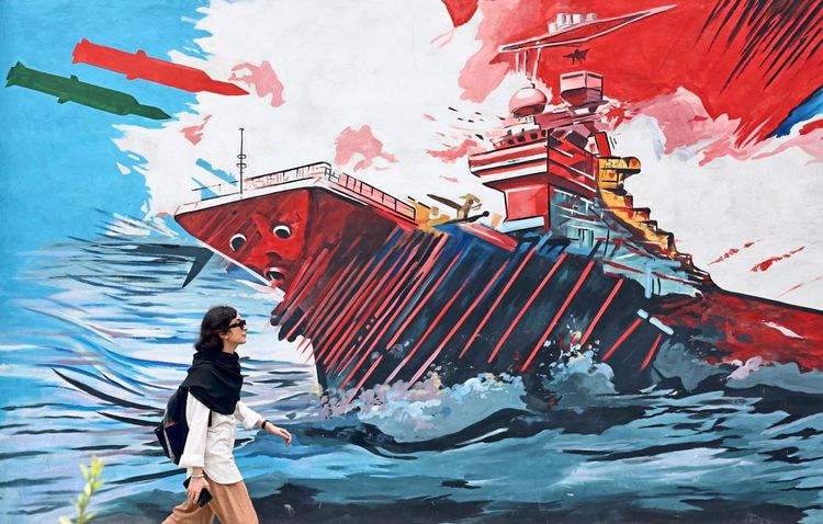 Eine Frau geht an einem Wandgemälde mit anti-amerikanischer Graffiti-Kunst in Teheran, Iran, vorbei. Das Graffiti zeigt ein großes Kriegsschiff in einer dramatischen Szene, umgeben von Wellen und farbintensiven, abstrakten Elementen.