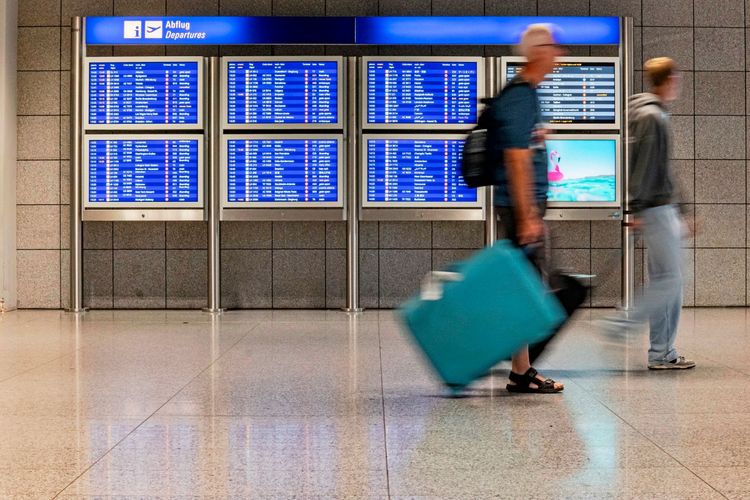 Zwei Passagiere gehen mit Gepäck an einer digitalen Abflugtafel im Frankfurter Flughafen vorbei. Einer der Reisenden zieht einen türkisfarbenen Rollkoffer. Im Hintergrund sind Flugverbindungen und eine touristische Werbeanzeige sichtbar.