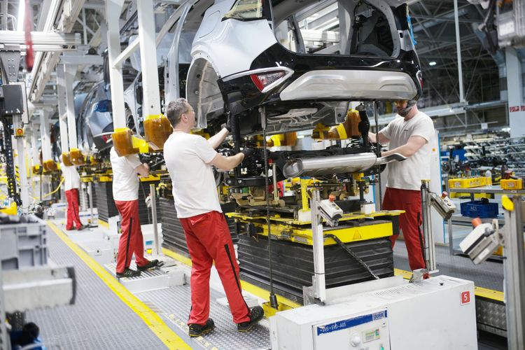 Arbeiter mit roten Hosen schrauben an einem halbfertigen Auto in einer Fabrikhalle.