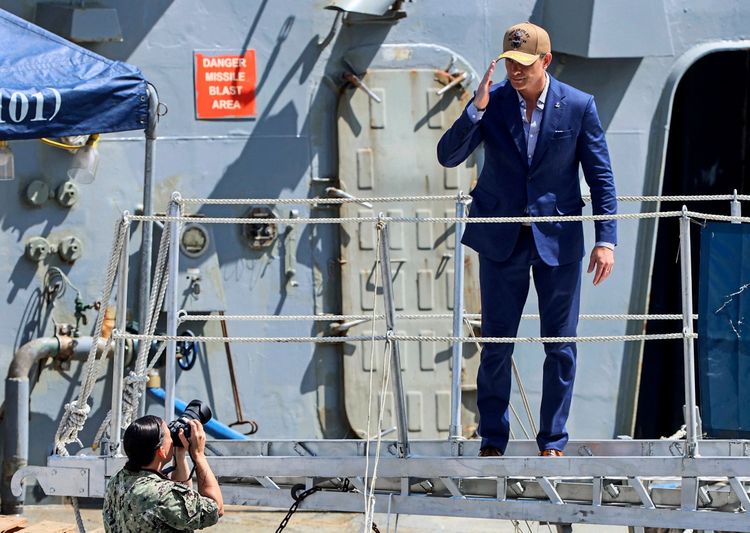 Ein Mann in einem blauen Anzug und einer Baseballkappe steht auf einem Schiffsdeck und salutiert. Im Hintergrund ist die Wand eines Schiffes mit dem Schild „Danger Missile Blast Area“ zu sehen. Im Vordergrund fotografiert eine Person in Militäruniform den salutierenden Mann.