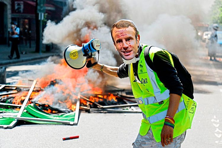 Plakate brennen und ein Mann steht in gelber Weste und einer Macron-Maske auf der Straße.