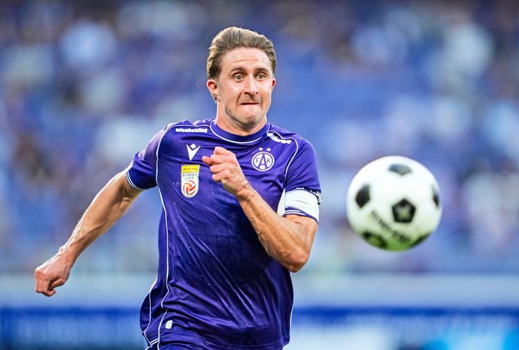 Ein Fußballspieler in einem violetten Trikot von FK Austria Wien läuft konzentriert einem Ball hinterher. Im Hintergrund ist ein unscharfes Stadion erkennbar.