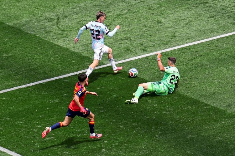 Ein Fußballspieler im weißen Trikot (Belgien, #22) läuft mit dem Ball am Liegen verteidigenden Torhüter im grünen Trikot (Spanien, #23) vorbei. Ein weiterer Spieler im roten Trikot (Spanien) rennt von hinten herbei. Die Szene spielt auf einem Fußballfeld.