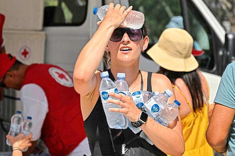 Eine Frau in Athen kühlt ihre Stirn mit einer Wasserflasche. In der anderen, der linken Hand hält sie fünf weitere Wasserflaschen.