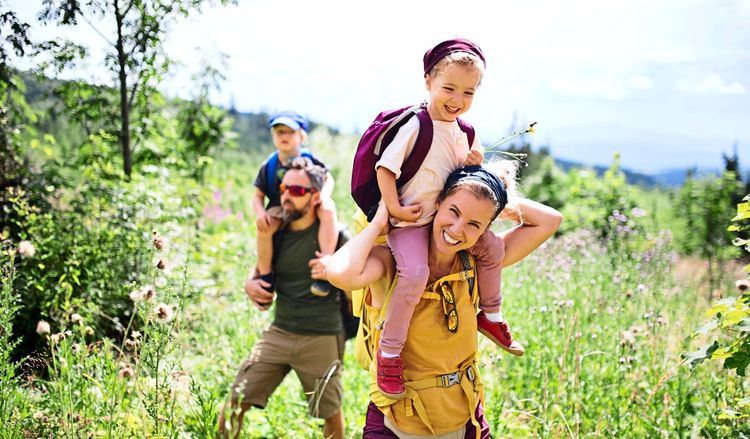 Eine Familie mit zwei kleinen Kindern wandert an einem sonnigen Sommertag durch eine grüne, blühende Landschaft. Ein Kind sitzt auf den Schultern der Mutter, das andere auf den Schultern des Vaters. Alle wirken fröhlich und genießen die Natur.