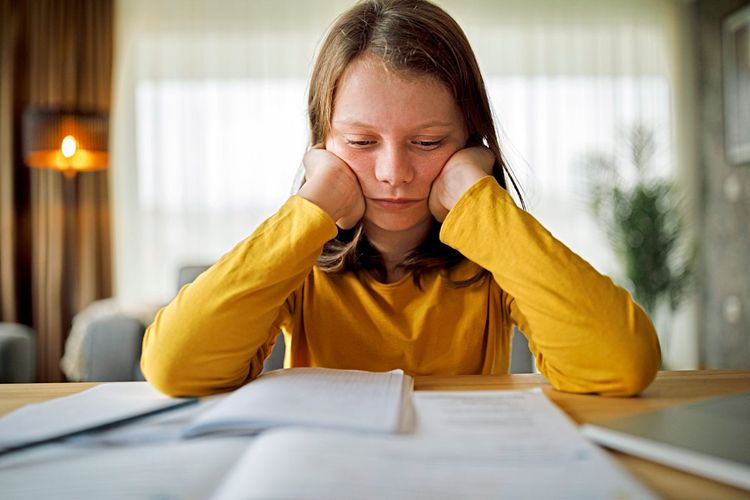 Mädchen in gelbem Shirt stützt ihren Kopf auf die Hände und blickt genervt auf die Hausaufgaben vor ihr 