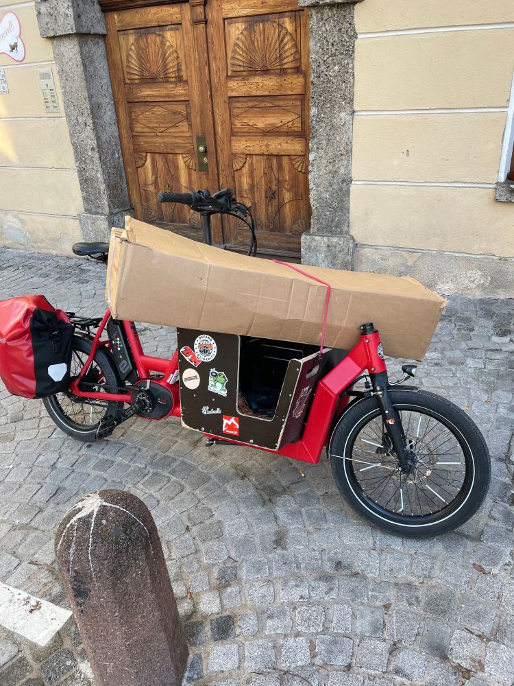 Ein lastenrad mit einem großen Karton, der auf der Lastenschale befestigt ist.