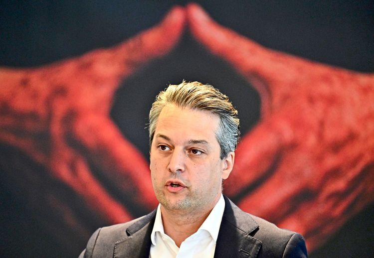 Ein Mann in Anzug und weißem Hemd spricht vor einem Hintergrund mit einem roten Motiv, das zwei Hände, die eine Raute bilden, zeigt.