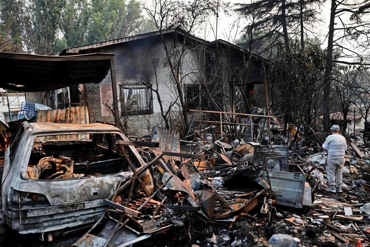 Ein vollständig abgebranntes Haus und ein zerstörtes Auto sind von Trümmern und Schrott umgeben. Ein Mann steht mit dem Rücken zur Kamera und betrachtet das Ausmaß der Zerstörung nach einem verheerenden Waldbrand in Pelayos de la Presa, westlich von Madrid, am 26. Juli 2026. Verkohlte Bäume und verbrannte Gegenstände sind sichtbar.