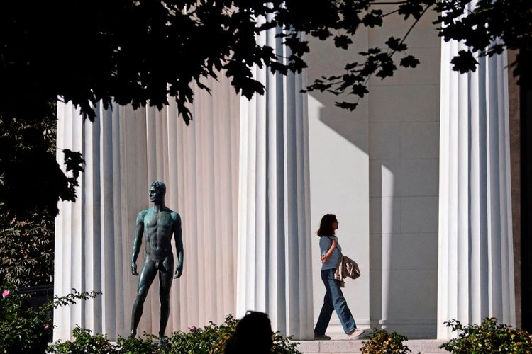 Eine Frau geht am neoklassizistischen Theseustempel im Volksgarten in Wien, Österreich, vorbei. Neben ihr steht eine bronzene Statue. Die Szene spielt sich bei Sonnenlicht ab, umgeben von Säulen und Pflanzen.
