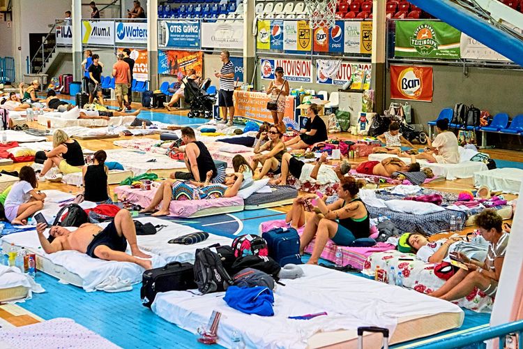 In einem umfunktionierten Sportstadium schlafen und sitzen etliche Touristinnen und Touristen auf Matratzen.