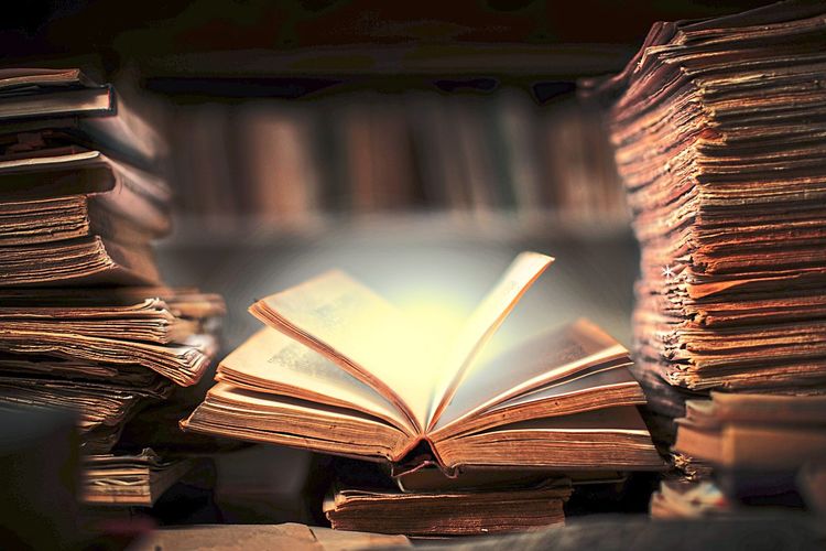 Ein vergilbtes Buch liegt geöffnet zwischen Stapeln alter Bücher, aus dem Buch scheint es zu leuchten