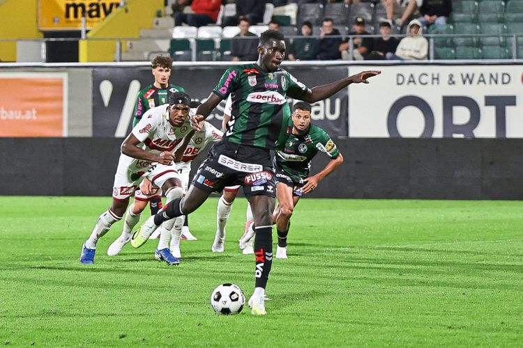 Ein Spieler des SV Ried (in schwarzem und grünem Trikot) kontrolliert den Ball während eines Spiels in der Admiral Bundesliga. Im Hintergrund sind weitere Spieler und Zuschauer auf der Tribüne zu sehen.