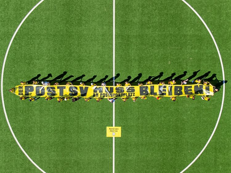 Luftaufnahme eines Fußballfeldes. Eine Gruppe Menschen hält ein großes gelbes Banner mit der Aufschrift: „DER POST SV MUSS AM POSTSPORTPLATZ BLEIBEN!“ Darunter ein kleines Plakat: „Rettet den POST SV“.