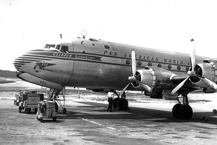 Schwarz-Weiß-Foto eines historischen Propellerflugzeugs der Pan American World Airways mit dem Namen „Clipper Endeavor“. Das Flugzeug steht auf einem Flughafen, im Vordergrund sind Transportwägen und ein Arbeiter sichtbar.